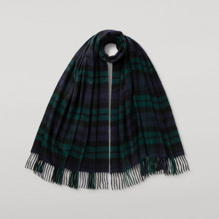 A dark green and black plaid scarf with fringe detailing, draped over a circular display stand against a light gray background.